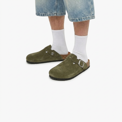 Green suede clogs worn with white socks and blue jeans on a white background