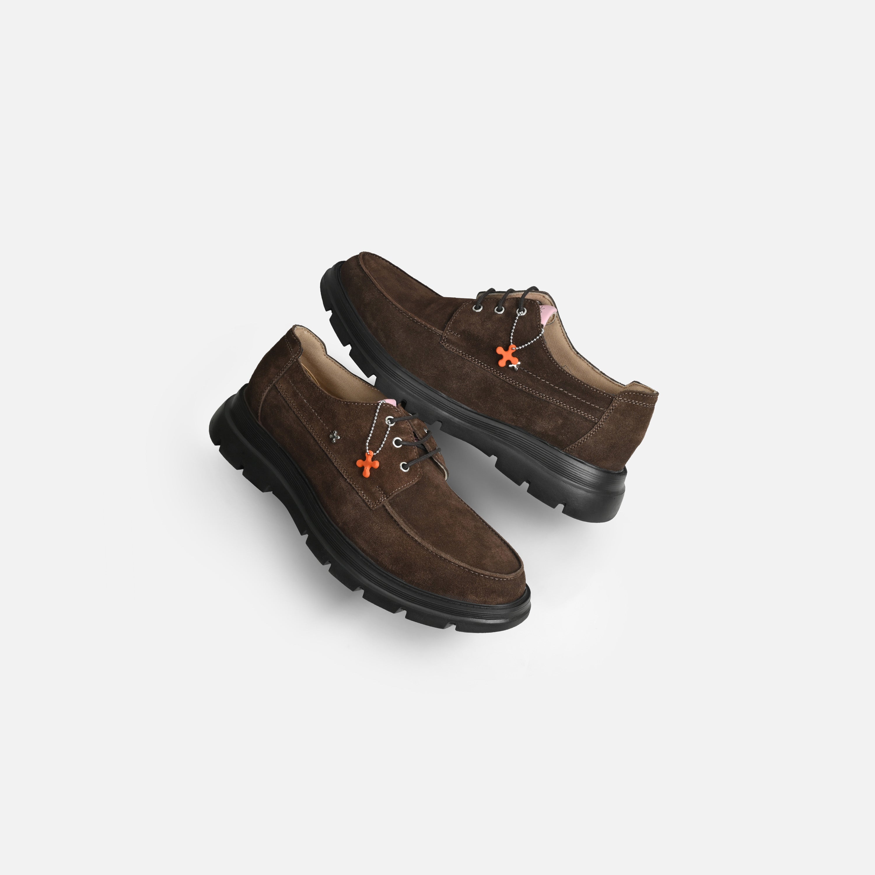 A pair of dark brown suede lace-up shoes with black rubber soles and small orange flower-shaped accents on the sides, displayed on a light gray background.