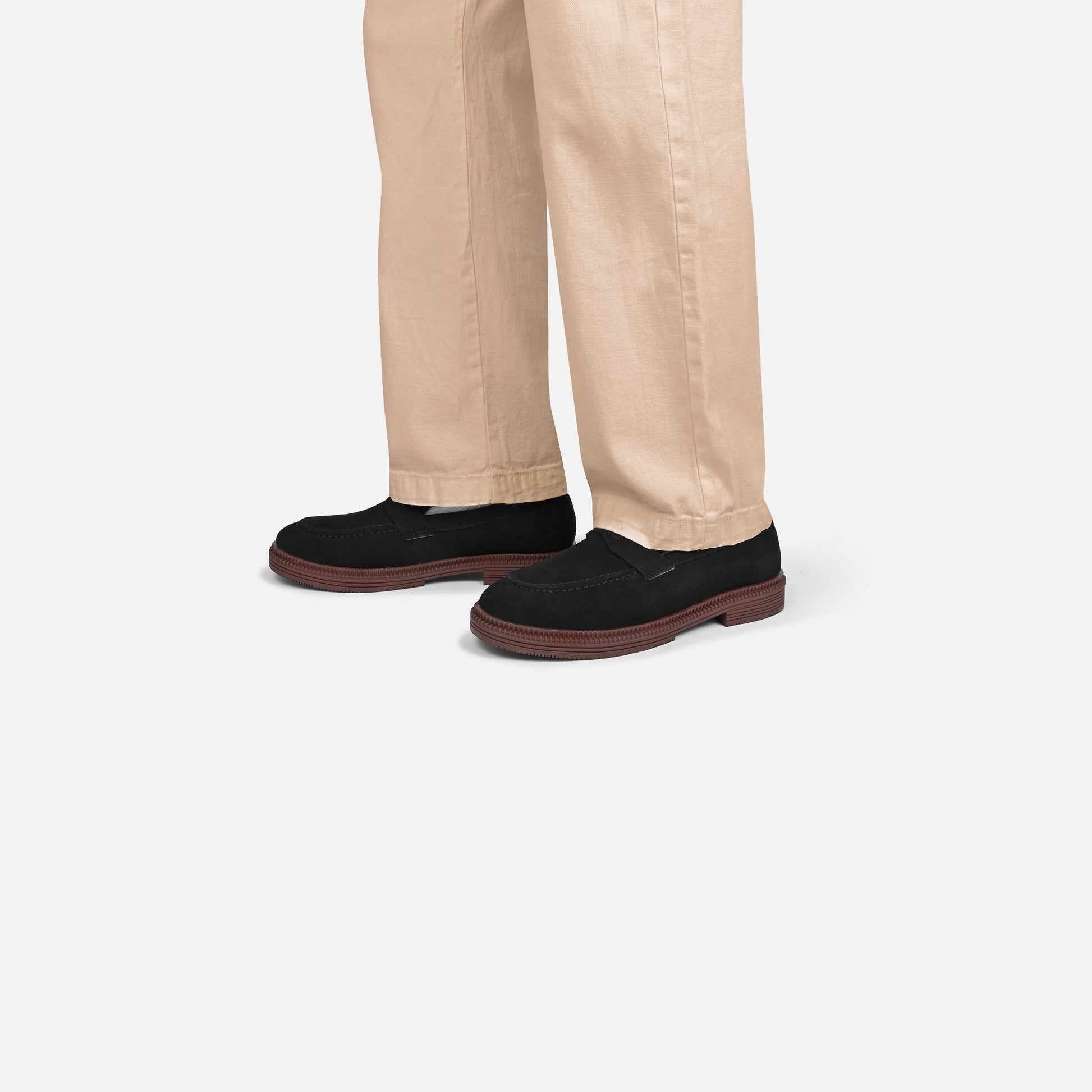 A person wearing beige trousers and black slip-on loafers with brown soles, shown from the knees down against a light gray studio background.