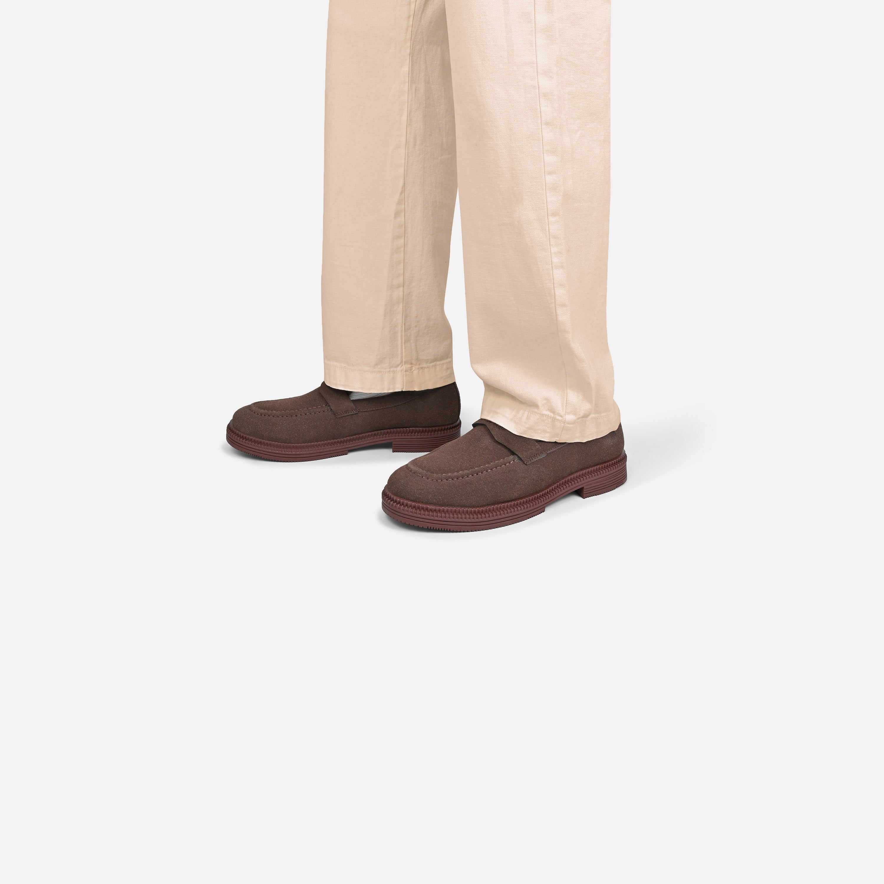 A person wearing beige trousers and dark brown slip-on shoes, shown from the calves down against a light gray studio background.