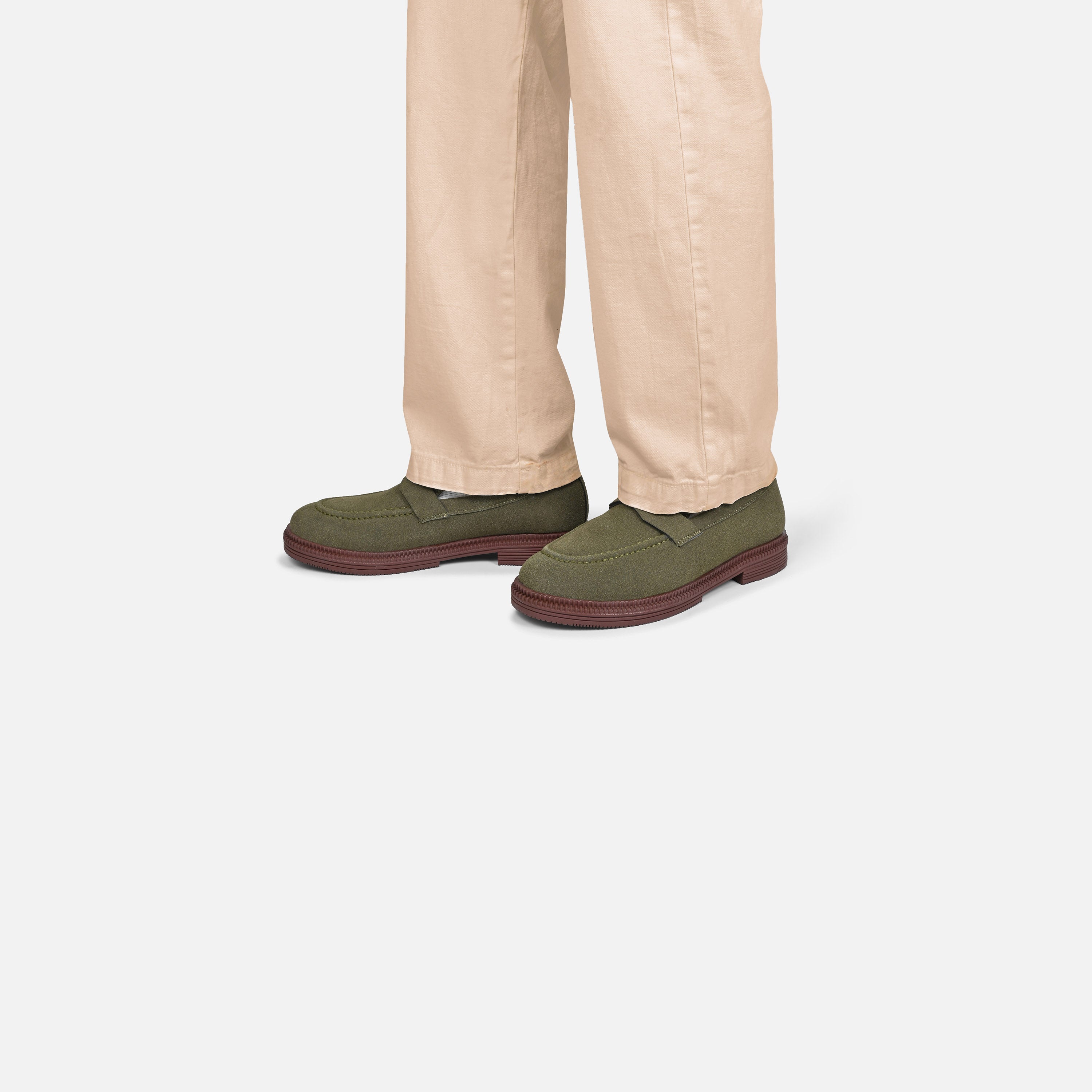 A person wearing beige trousers and olive green slip-on shoes with dark brown soles, shown from the knees down against a light gray studio background.