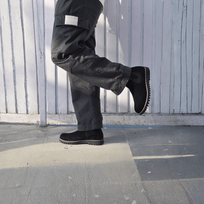 Person wearing black work boots and pants on a tiled floor with a wooden panel background