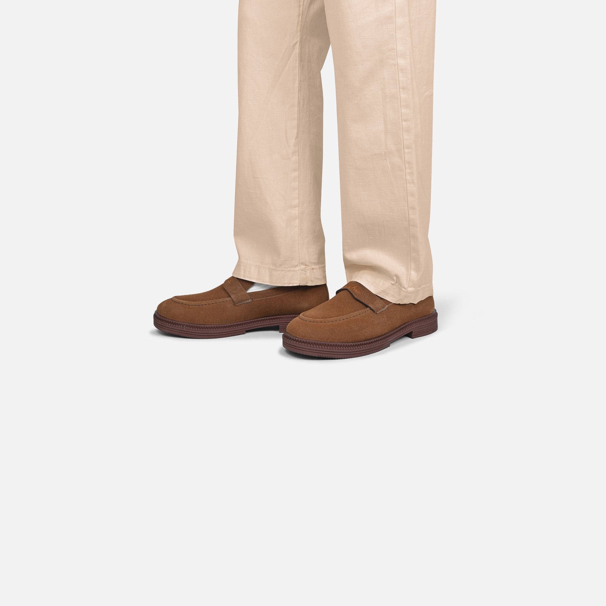 A person wearing beige trousers and brown suede slip-on loafers, shown from the knees down against a light gray studio background.
