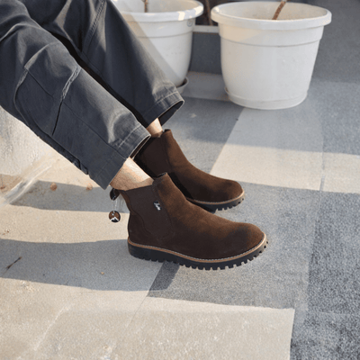 Brown ankle boots worn by a person sitting on steps with potted plants in the background.