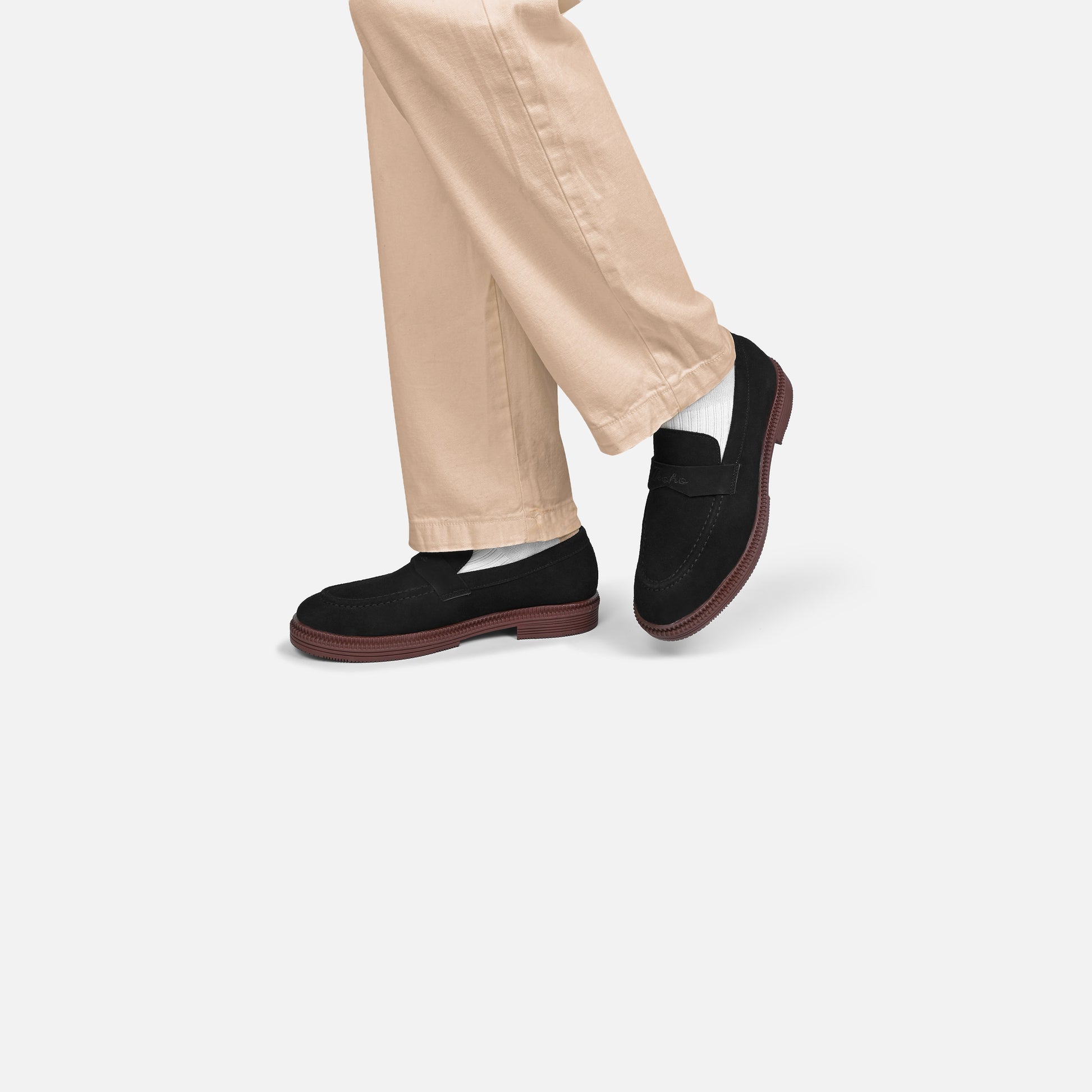 A person wearing beige trousers and black slip-on loafers with white socks, shown from the knees down against a light gray studio background.