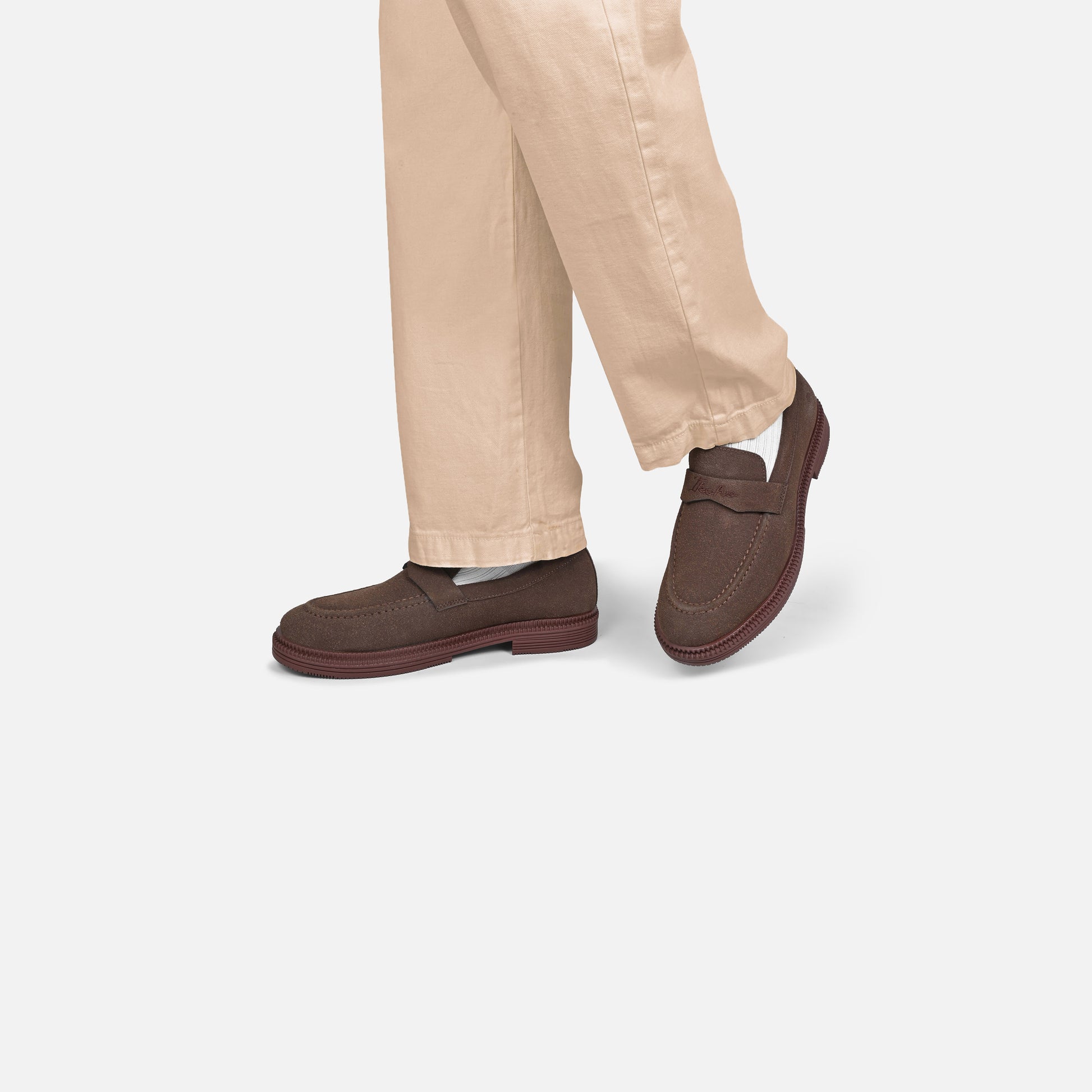 A person wearing dark brown leather slip-on loafers with beige pants, shown from the knees down against a light gray studio background.