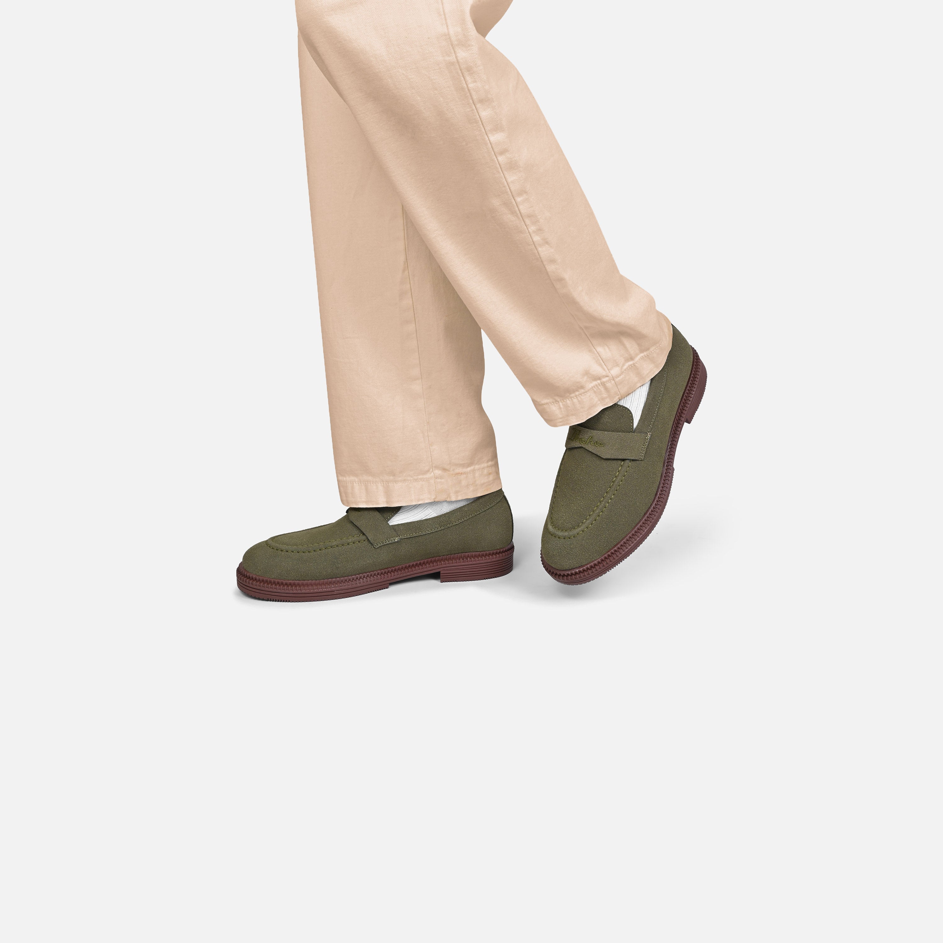 A person wearing olive green slip-on loafers with dark brown soles, paired with light beige trousers, shown from the knees down against a light gray studio background.