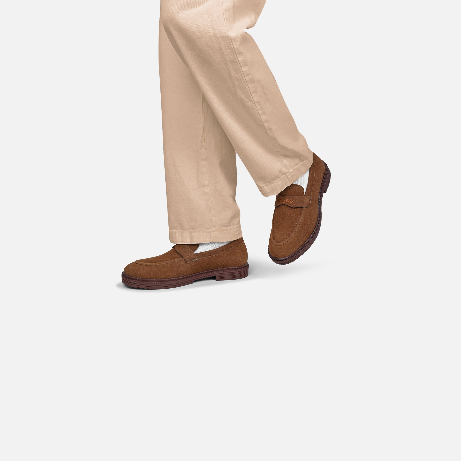 A person wearing tan chinos and brown suede penny loafers, shown from the knees down against a light gray studio background.