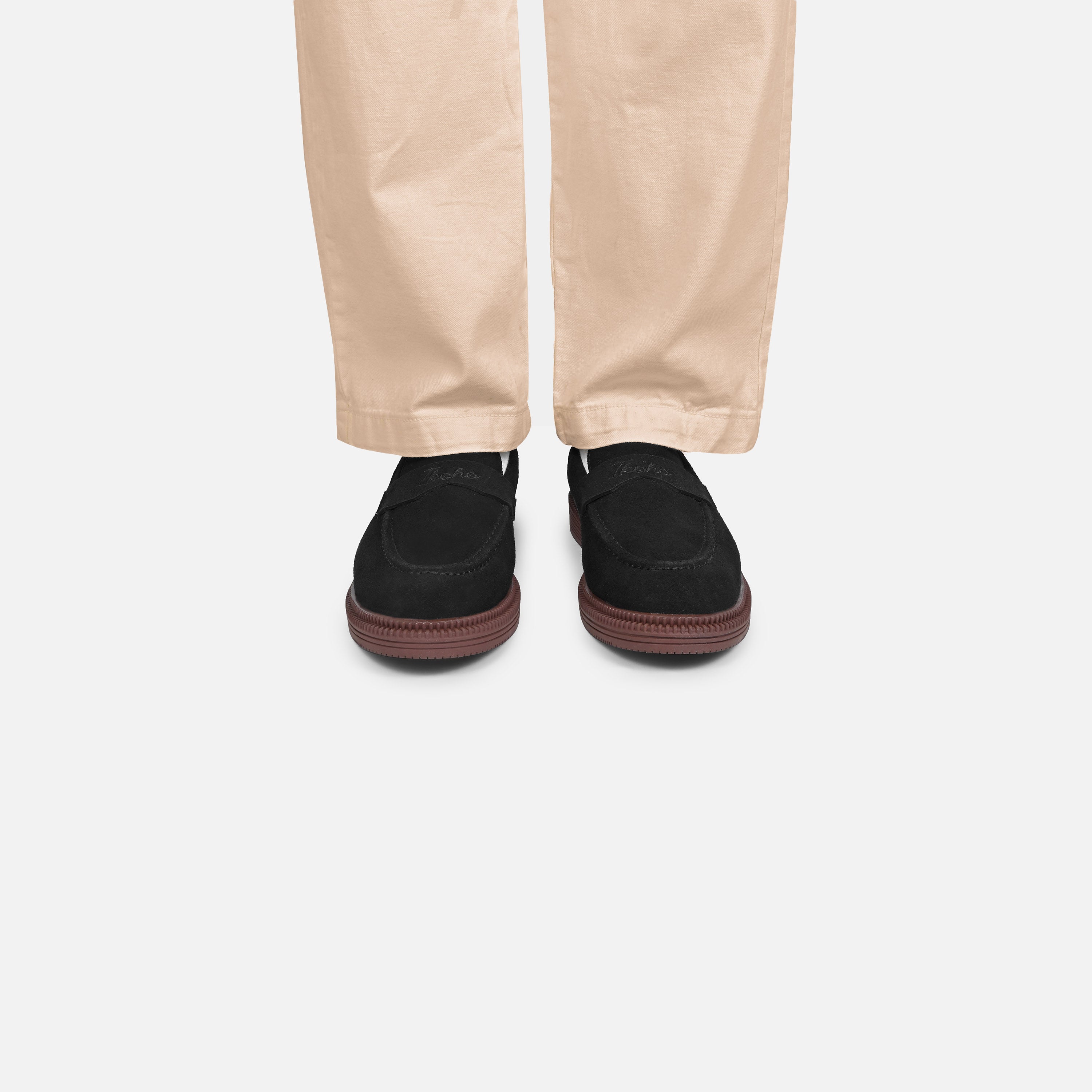 A person wearing beige trousers and black slip-on loafers with dark brown soles, shown from the feet up against a light gray background.