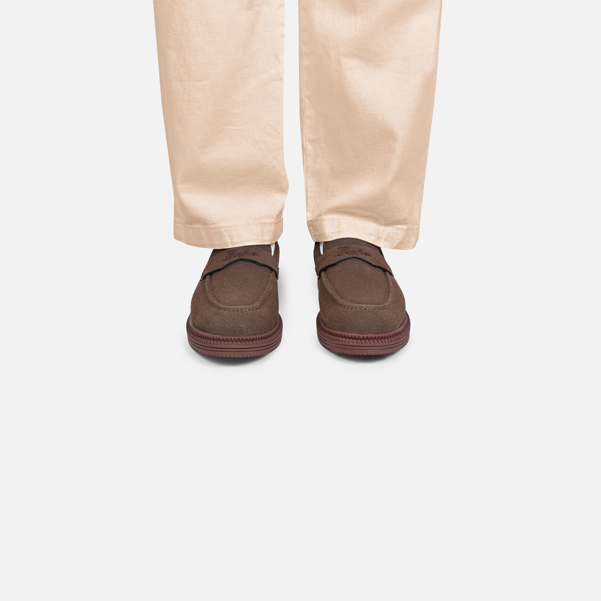 A pair of dark brown slip-on loafers worn with light beige trousers, shown from the back against a light gray background.
