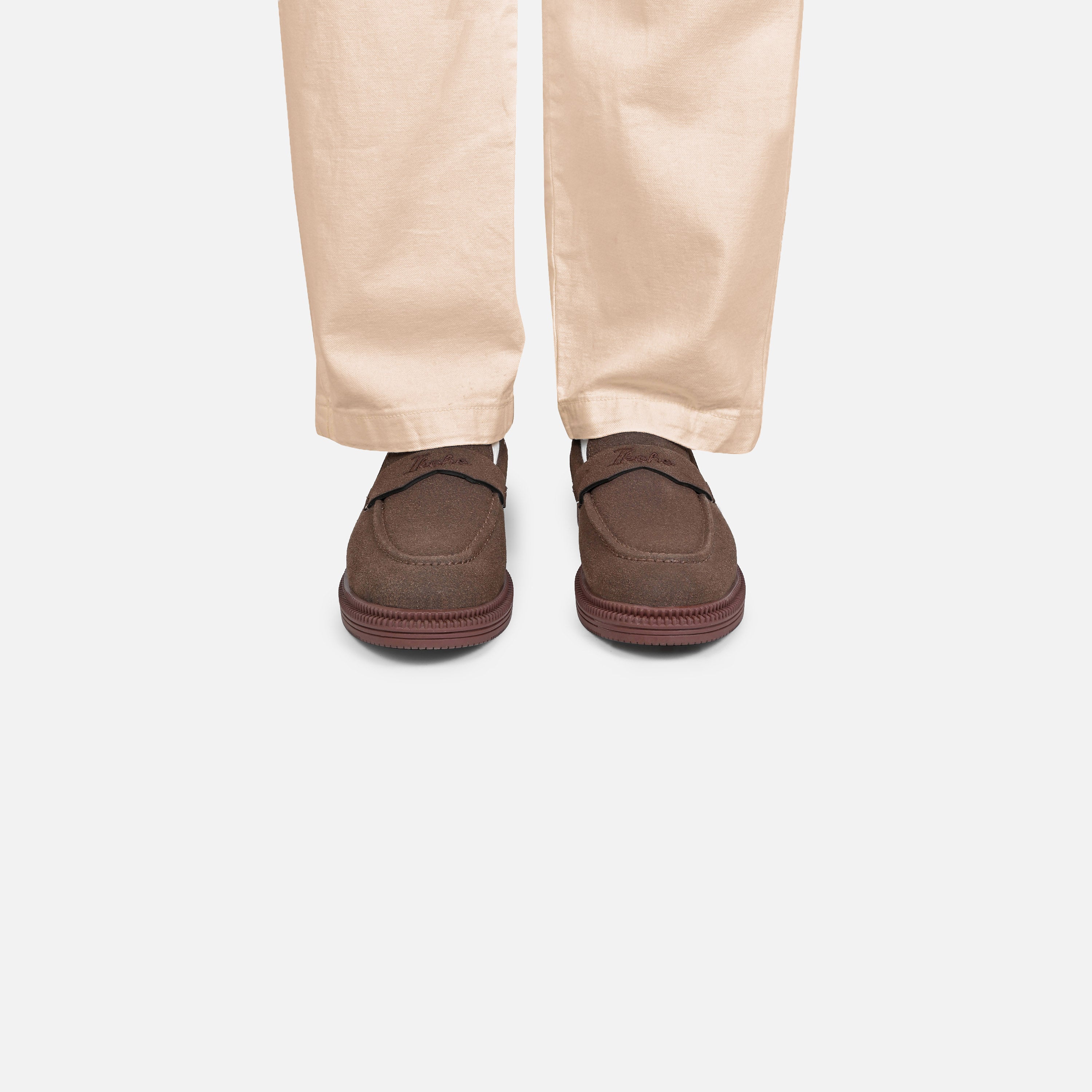 A pair of dark brown slip-on loafers worn with light beige trousers, shown from the back against a light gray background.