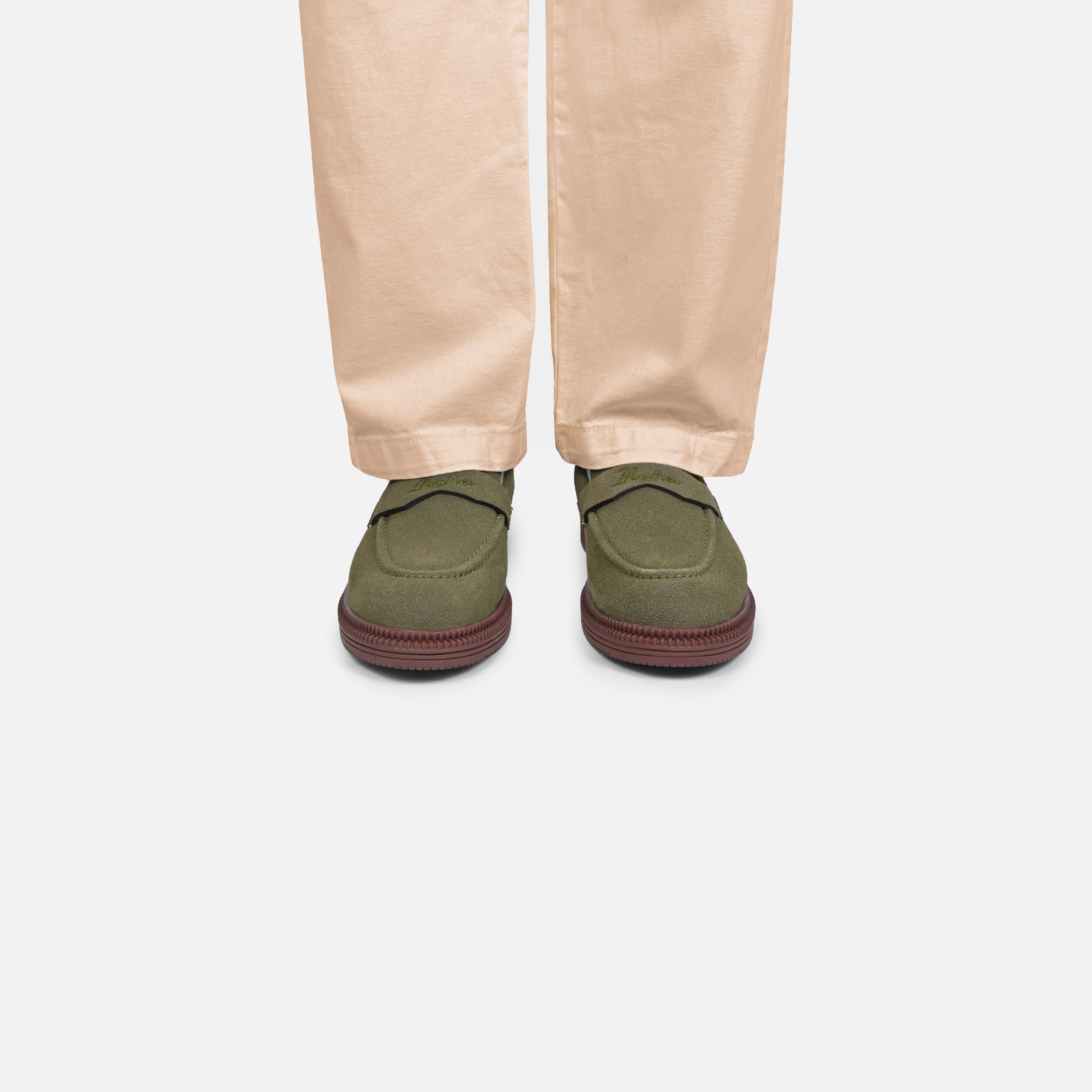 A person's lower legs wearing beige trousers and olive green slip-on shoes with dark reddish-brown soles, shown from the back against a light gray background.