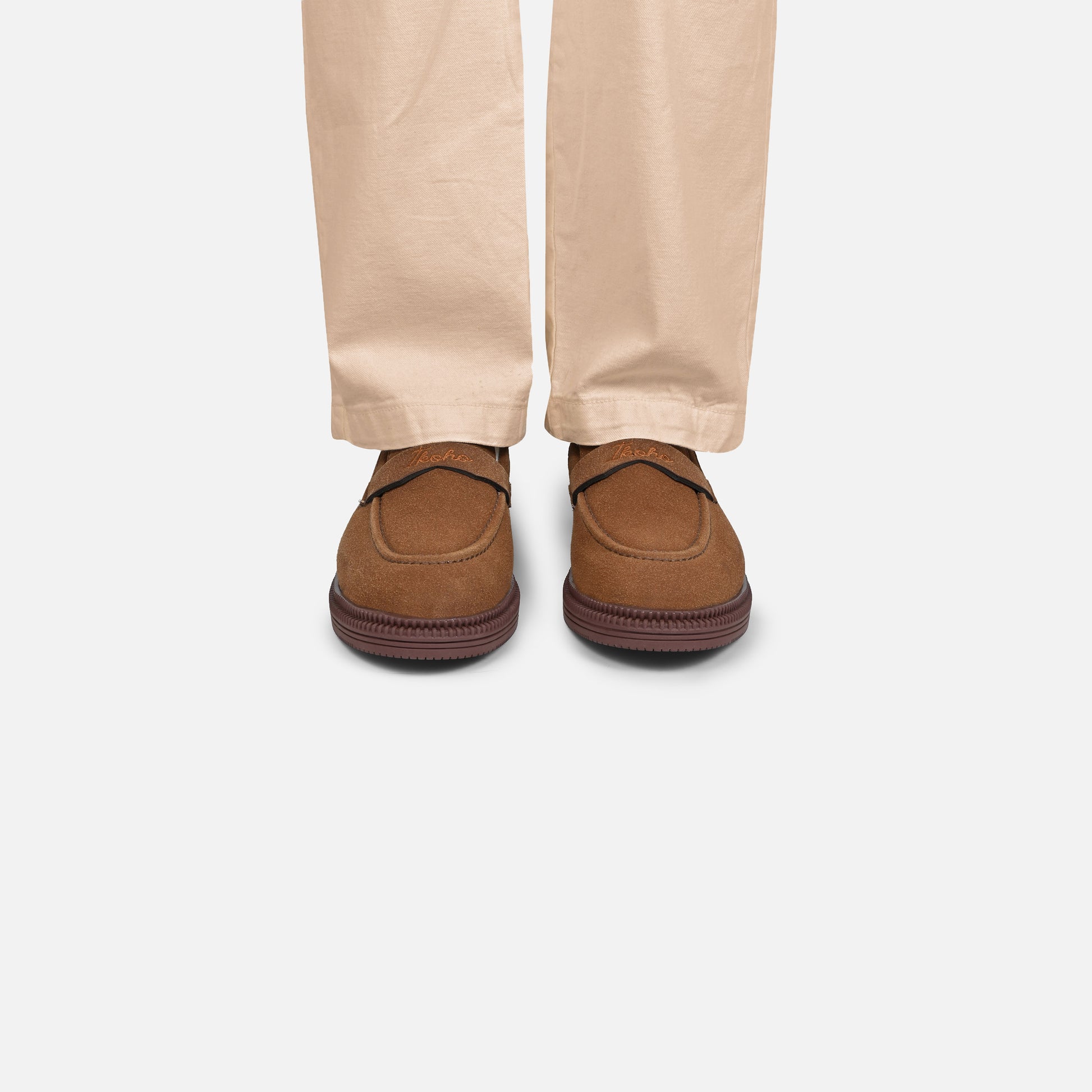 A person wearing tan pants and brown suede slip-on loafers, shown from the feet up against a light gray studio background.