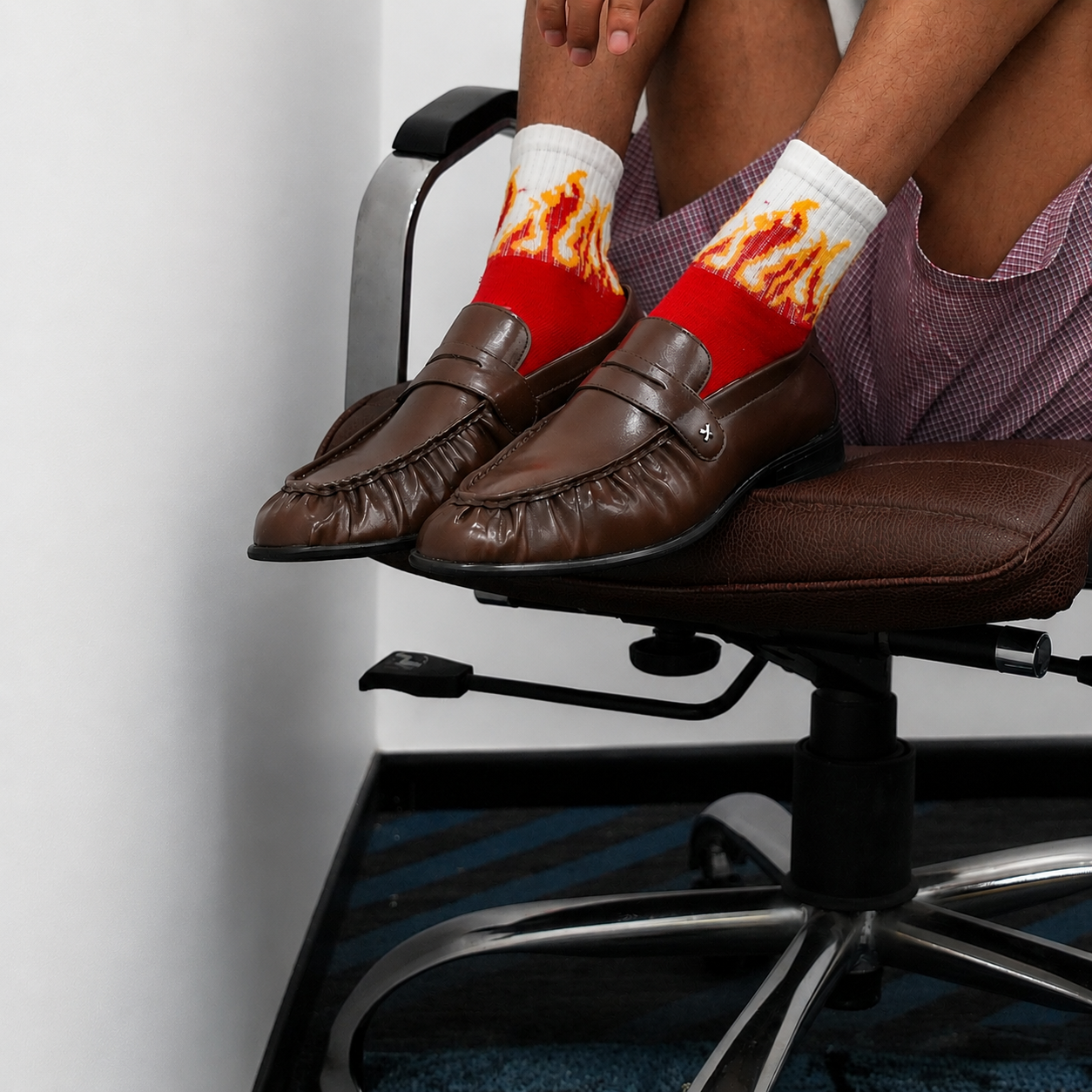 A person sitting in a brown office chair with their legs crossed, wearing brown leather loafers and red crew socks with a yellow and orange flame pattern on a white cuff.
