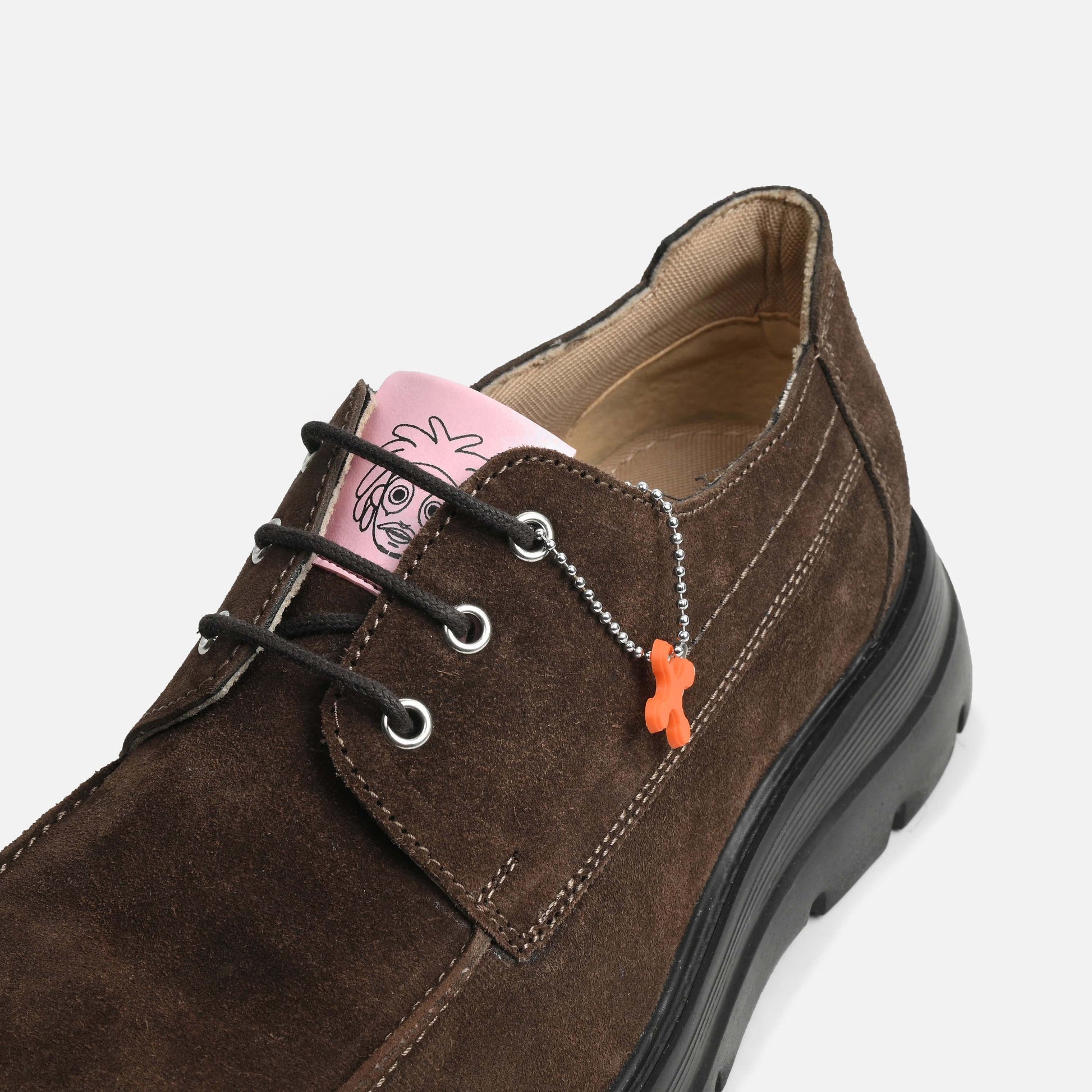 A close-up of a dark brown suede lace-up shoe with black laces, silver eyelets, a black sole, and a small orange dog-shaped charm on a silver chain. A pink paper tag is visible inside the collar.