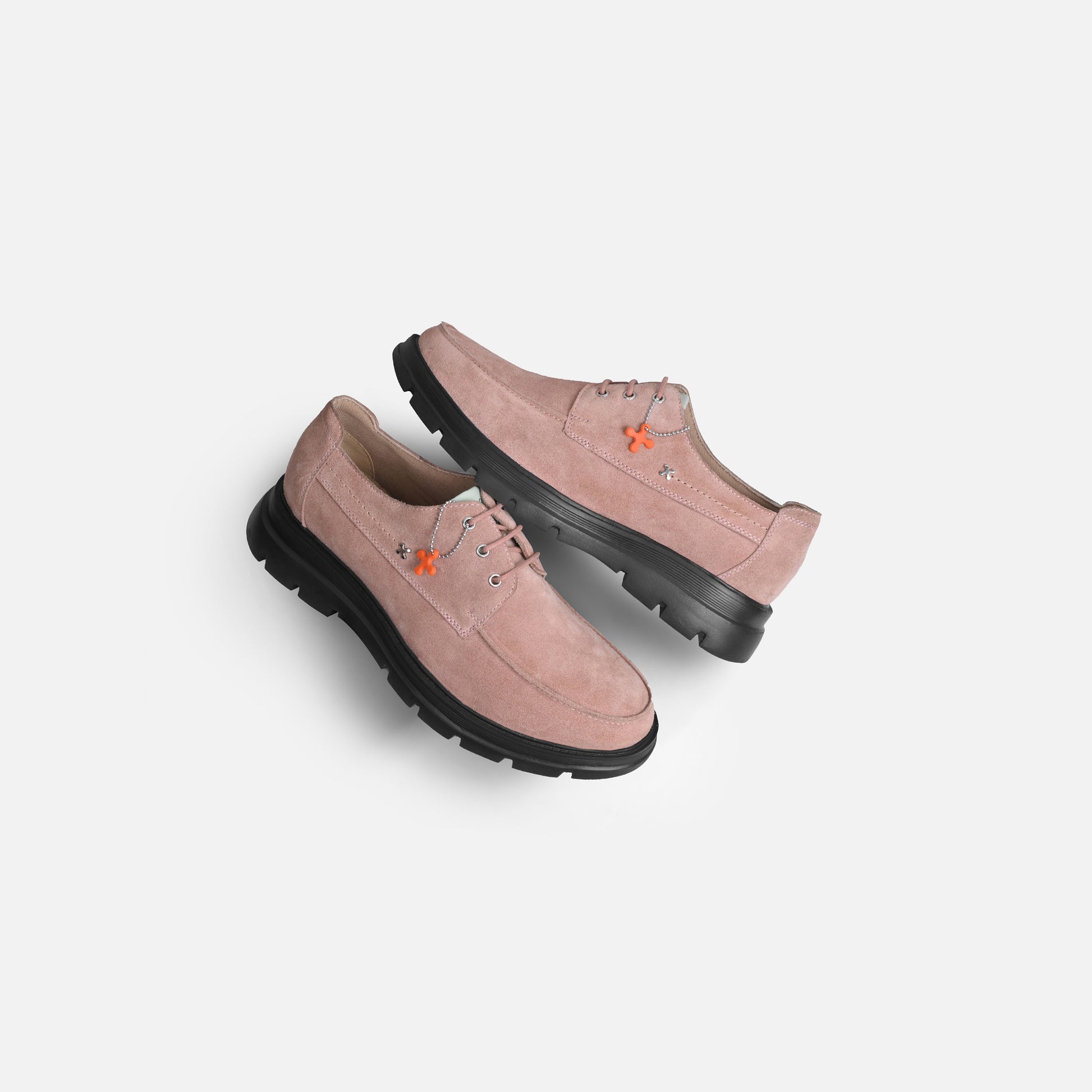 A pair of dusty pink suede lace-up shoes with chunky black soles and small orange embroidered details, shown on a light gray background.