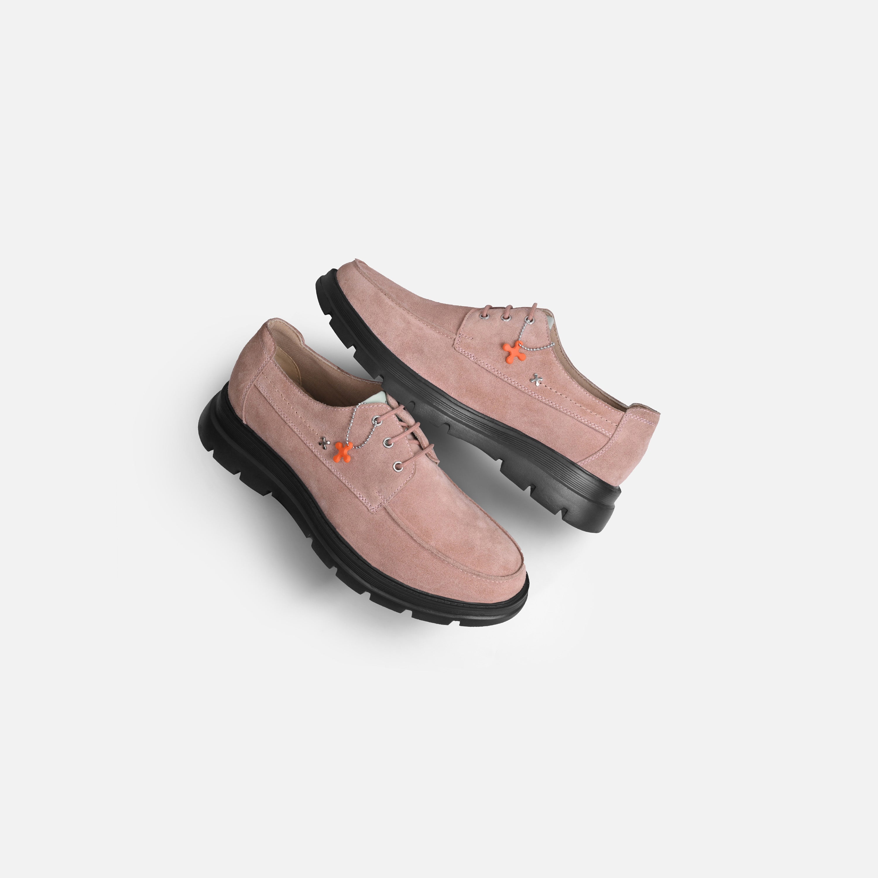 A pair of dusty pink suede lace-up shoes with chunky black soles and small orange embroidered details, shown on a light gray background.