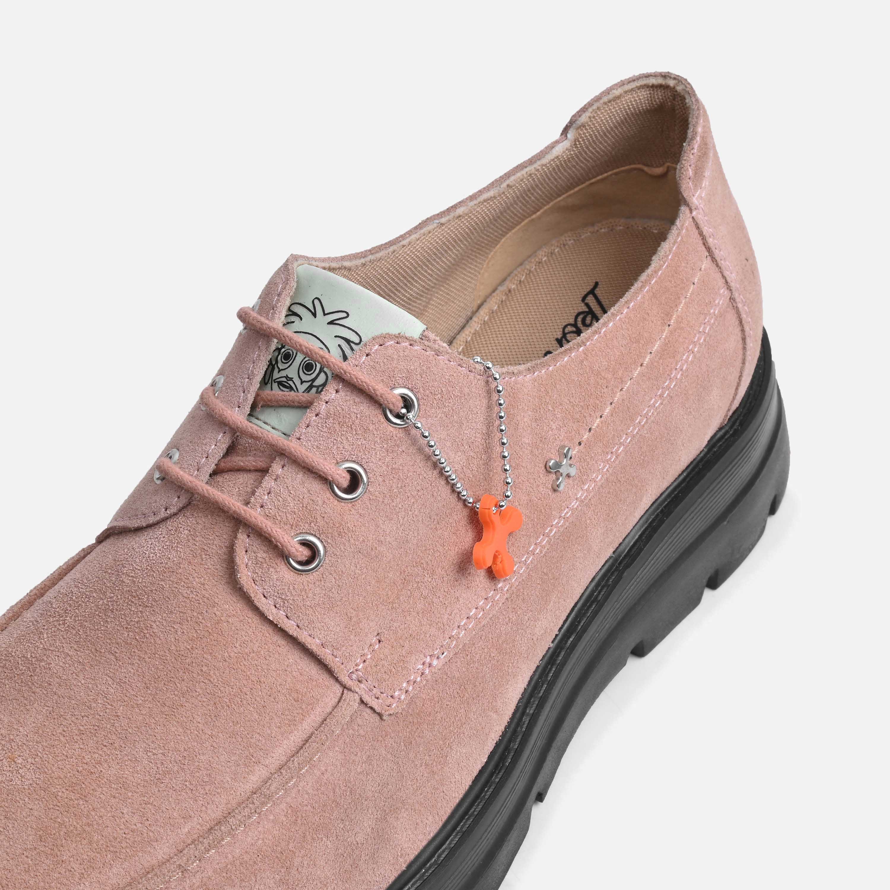 A dusty pink suede lace-up shoe with a chunky black sole, featuring a silver chain accessory with an orange cross-shaped charm hanging from the eyelet.
