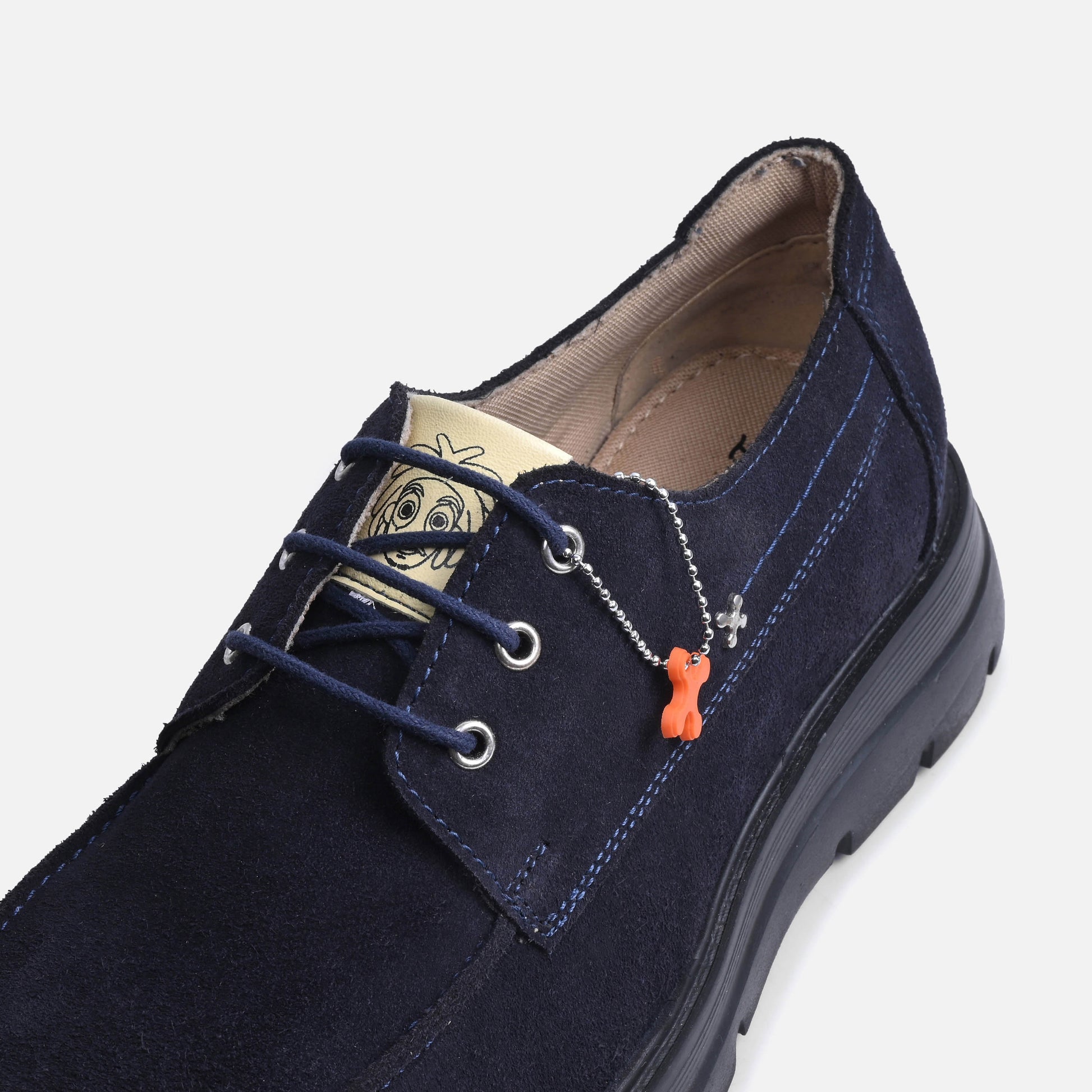 A navy blue suede lace-up shoe with a chunky black sole, featuring a decorative silver chain with an orange bone-shaped charm and a beige brand tag on the tongue.