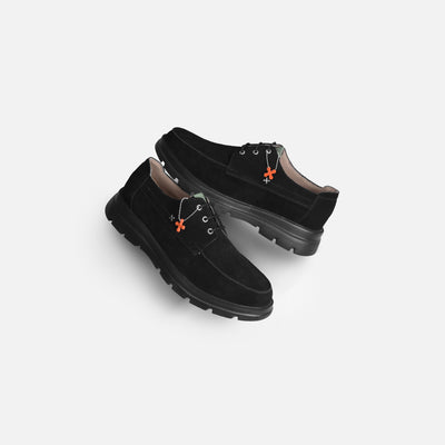 A pair of black suede lace-up shoes with chunky black soles and small orange decorative accents on the eyelets, displayed on a light gray background.