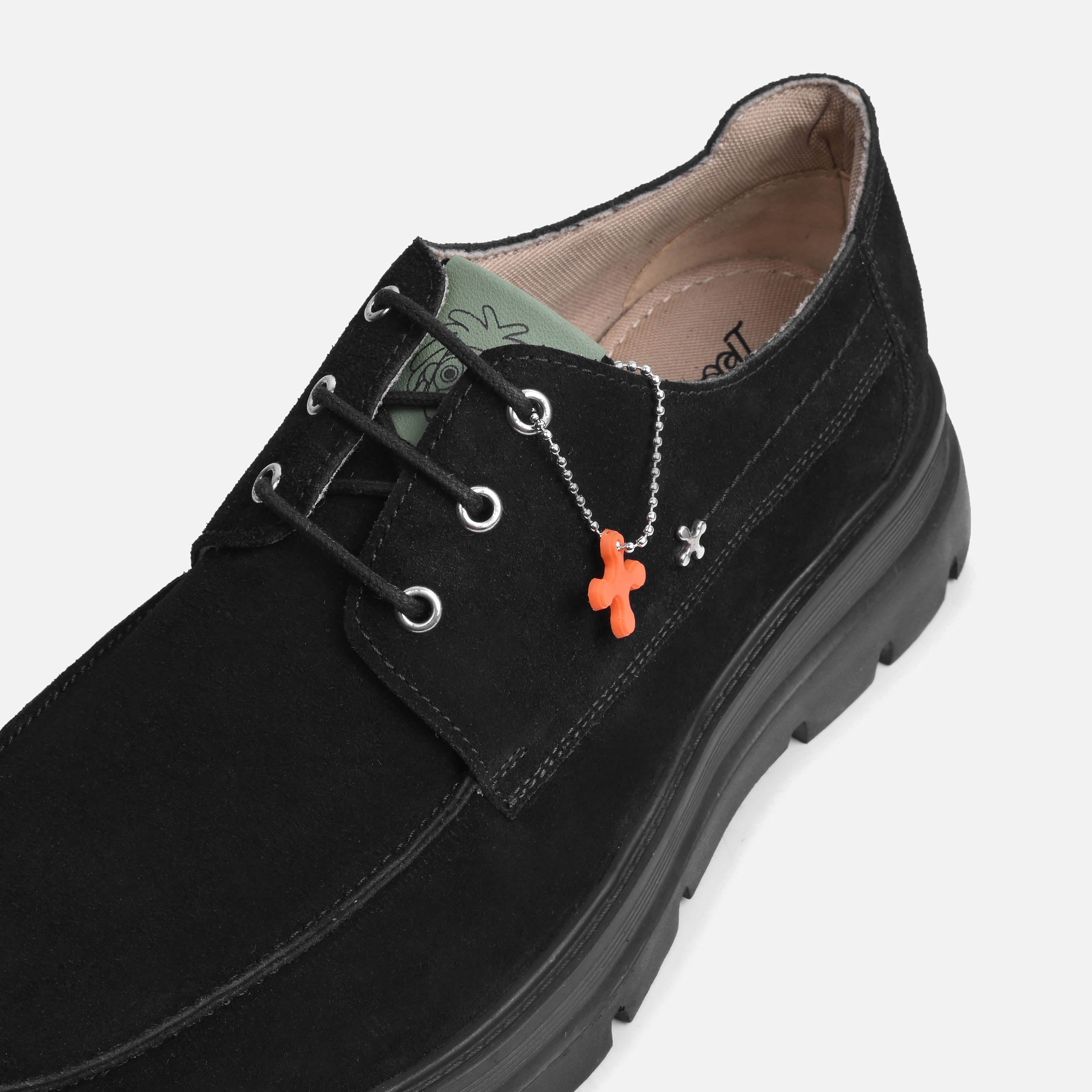 A black suede lace-up shoe with a chunky black sole, featuring a silver chain necklace with an orange cross charm hanging from the side eyelet.