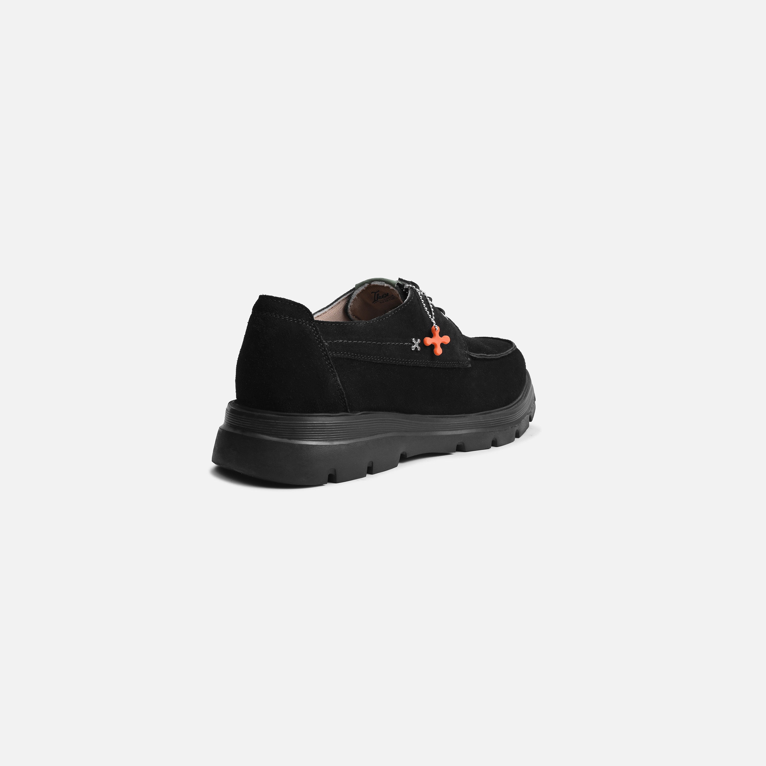A black suede loafer-style shoe with a chunky black sole and a small orange cross detail on the side, shown from a rear three-quarter angle against a light gray background.