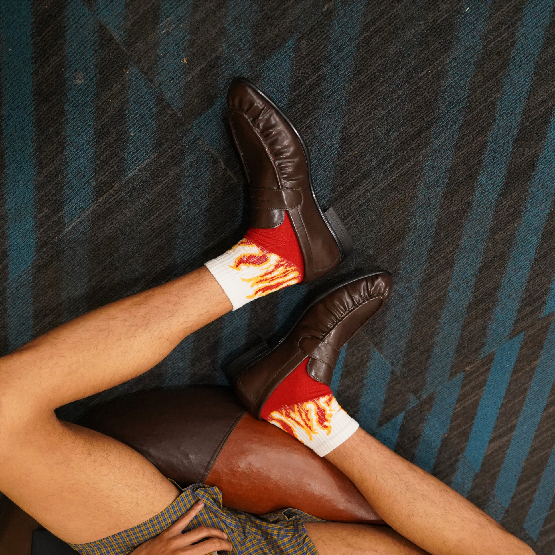 A person reclining on a blue and gray striped carpet, wearing dark brown leather loafers and white crew socks with a red and orange flame print.