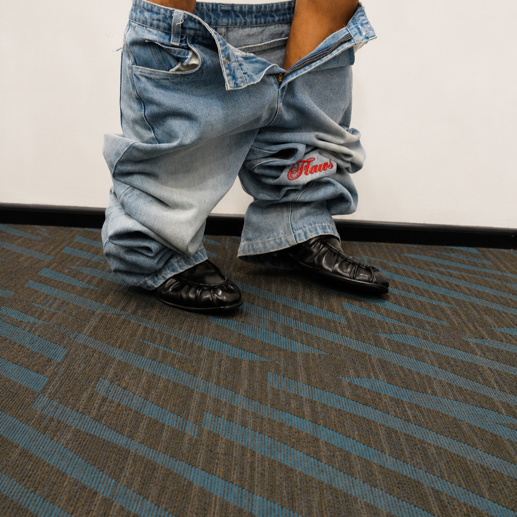 A person wearing baggy light blue denim jeans and black shoes, standing on a blue and gray patterned carpet against a plain white wall.