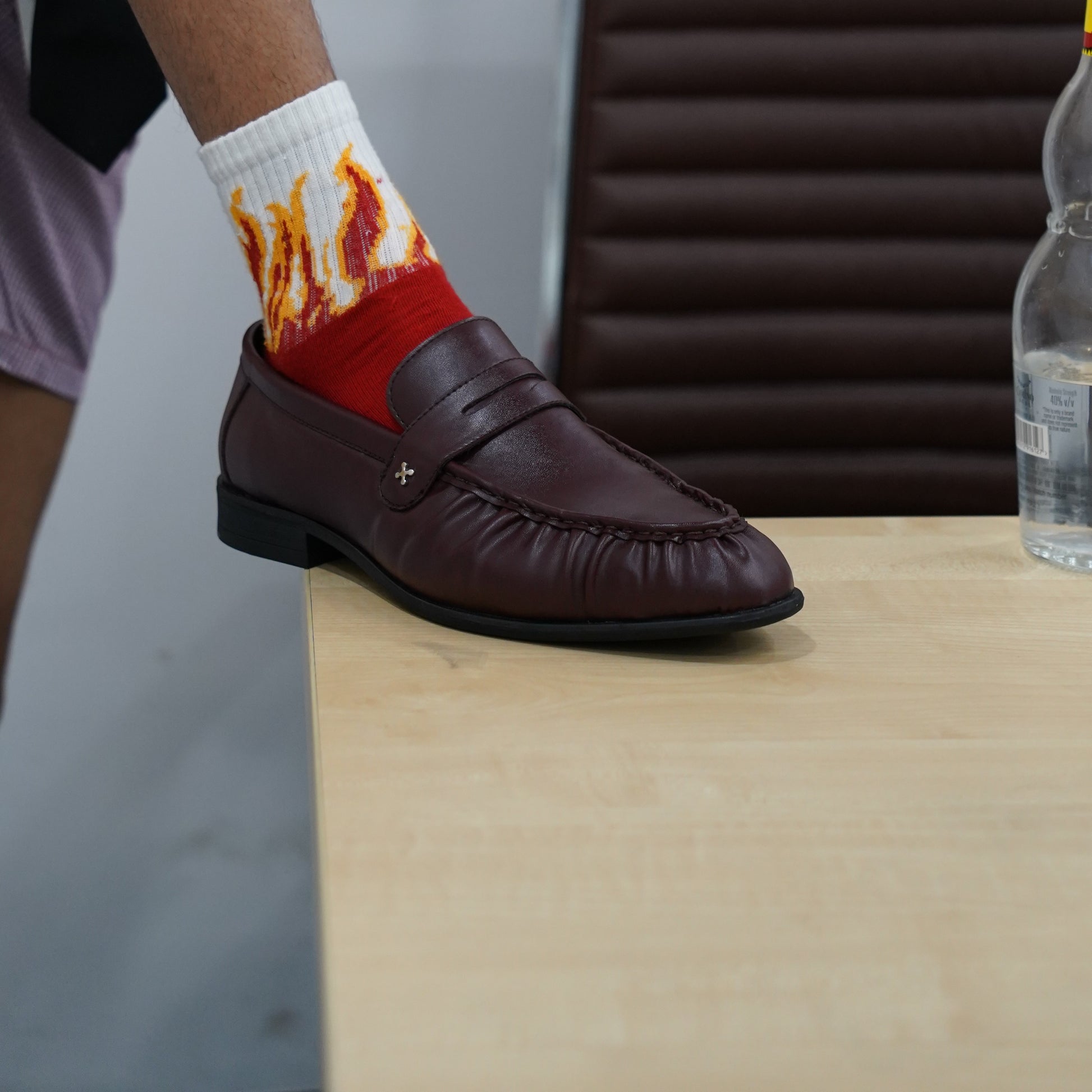 A person sitting on a brown leather chair wearing brown leather penny loafers and white crew socks with a red and orange flame print, set on a blue and gray patterned carpet.