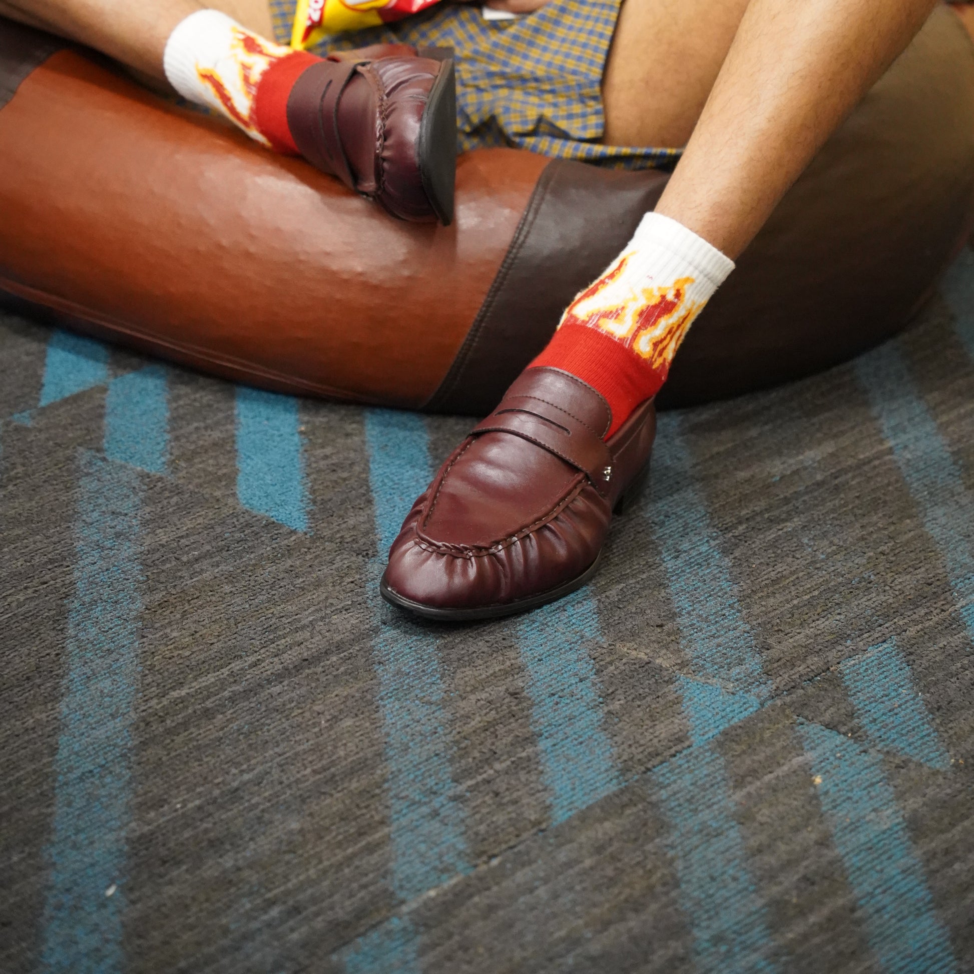 A person sitting on a brown leather chair wearing brown leather penny loafers and white crew socks with a red and orange flame print, set on a blue and gray patterned carpet.
